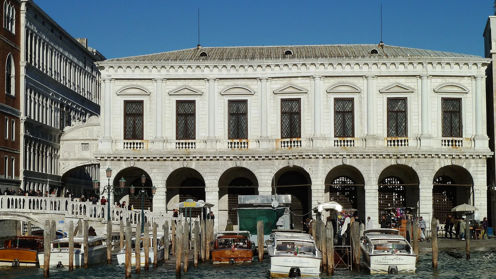 The white arcaded front of Venice's Prisons Palace seen from the lagoon, with the Bridge of Sighs at its left edge and water taxis moored in front