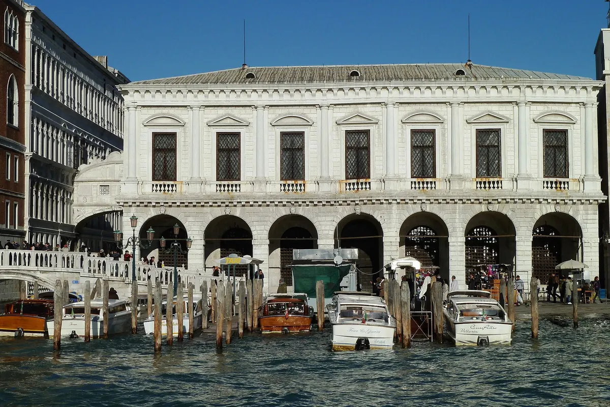 The white arcaded front of the New Prisons seen from the lagoon side, with water taxis moored in front and the Bridge of Sighs at the left edge.